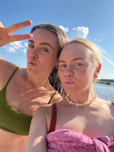 Two women are taking a selfie outdoors, with a bright blue sky and fluffy white clouds overhead. The woman on the left is wearing a green bikini top and has dark hair. She is holding up her hand in a peace sign and puckering her lips. The woman on the right has blonde hair and is wearing a seashell necklace and a pink towel or sarong. She is also puckering her lips. They appear to be near a body of water, possibly a river or lake, with trees and vegetation visible in the distance. The lighting suggests it is a sunny day. This scene is located in the city of Ust'ye, Russia.
