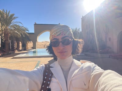 A woman is depicted taking a selfie outdoors at a desert resort in Rissani, Morocco, under bright sunlight, which produces a prominent lens flare in the upper right quadrant. The subject, centered in the foreground, has short, dark, curly hair and is wearing dark oval sunglasses, a multi-colored patterned headscarf (predominantly green, yellow, and red), a light-colored turtleneck sweater, and a cream-colored jacket with two breast pockets. A brown shoulder bag adorned with silver studs is visible over her right shoulder. Her right arm, extended to capture the image, is partially visible on the right.

The background features a long, narrow swimming pool with clear blue water. Along the left side of the pool, several palm trees stand alongside lounge chairs equipped with red cushions and white parasols, situated under a shaded colonnade with pillars. A large arched entryway in the mid-ground frames a view of extensive sand dunes, characteristic of the Sahara Desert. To the right, a multi-story building constructed from earthen-toned materials exhibits traditional Moroccan architectural elements, including stylized cactus-like decorative motifs on its facade. The ground area surrounding the pool is paved with light-colored tiles.