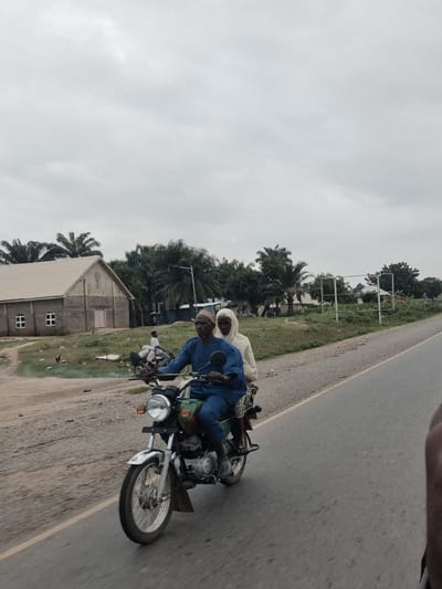 The image depicts a street scene in a rural area, likely a village, in Nigeria. The foreground is dominated by a motorcycle carrying two individuals. The driver, an adult male, wears a blue traditional outfit and a patterned cap. He is looking forward, seemingly focused on the road. Seated behind him is a passenger, an adult female, who is covered by a light-colored headscarf. They are both positioned on the motorcycle, suggesting they are traveling.

The motorcycle itself is a common model, with a visible headlight, engine, and wheels. It appears to be moving on a paved road with a yellow line marking the edge.

In the background, there are several structures and natural elements. A building resembling a church, constructed with grey blocks and featuring a prominent cross, is visible on the left. It has a sloped, metallic roof and two small, square windows. The area around the church appears to be a dirt path or unpaved ground, with some sparse vegetation and a few chickens visible.

Further in the background, there are numerous palm trees and other dense foliage, characteristic of a tropical environment. A metal billboard frame stands empty in the middle ground, suggesting the presence of advertising. The sky is overcast with a light gray hue, indicating a cloudy day. The overall scene suggests a typical day in a Nigerian village, with people engaged in daily transportation.