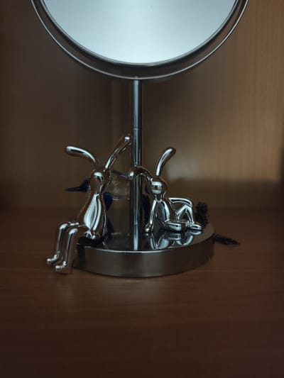 Two chrome-finished rabbit figurines are positioned on a wooden surface in front of a round mirror on a stand. The figurines appear to be sitting, with one slightly larger than the other. A dark, possibly black, cord or chain is visible trailing from the base of the mirror stand. The background is a plain, light brown surface, likely a shelf or wall. The lighting suggests an indoor setting, possibly a dressing table or vanity. No people are present in the image. This scene is not located in Krasnokamensk, Russia.