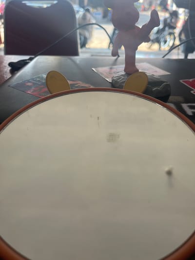 The image shows a close-up of a large, round, pale object with a brown rim, possibly a drumhead or a cymbal. In the background, a blurry figure of a person is visible, with what appears to be a toy or doll with yellow feet and arms in the foreground. The figure's hand is resting on the pale surface. The background also shows other objects, including what looks like a dark red object on the left, possibly a chair or a bag, and a window with reflections of lights and possibly trees. The overall setting appears to be indoors, with a somewhat cluttered environment. There is no clear indication of the time of day or weather. The image has a shallow depth of field, with the foreground and the central object in focus, while the background is blurred. Some faint, unreadable text is visible on the pale surface.