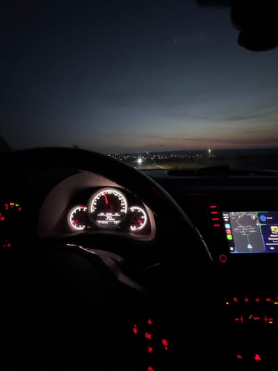The image depicts the interior of a moving vehicle, specifically from the driver's perspective, looking forward through the windshield at dusk or early night. The location is specified as Kumanovo, Bulgaria.
The foreground is dominated by the vehicle's dark steering wheel and dashboard. The instrument cluster is illuminated, showing a speedometer with the needle indicating 70 units (likely km/h). Below the speedometer, a digital display reads "18:45" (time), "125.0 km" (trip distance), and "5.5°C" (temperature). A tachometer on the left shows approximately 2000 RPM, and a fuel gauge on the right indicates approximately half full. Red warning lights are visible on the left of the cluster.
To the right of the steering wheel, an illuminated infotainment system displays a navigation map with a blue route and a blue directional arrow. Partially visible music information includes "About Time." Below the infotainment screen, various buttons are illuminated with red lights.
Through the windshield, a dark, open road is visible leading towards a horizon dotted with numerous small, illuminated points, characteristic of distant city lights, presumably Kumanovo. The sky above transitions from a dark blue overhead to a lighter, reddish-orange hue at the horizon, indicative of twilight conditions. Headlights from an oncoming vehicle are faintly visible in the distance. No individuals are directly visible in the frame, though the presence of an active vehicle interior implies a driver.