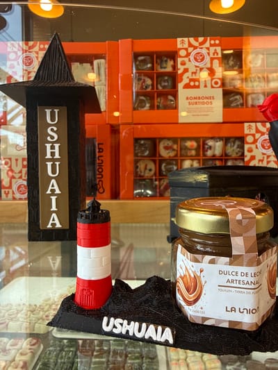 The image displays a clear glass display case featuring confectionery and souvenir items within a retail establishment. In the foreground, from left to right, are three primary objects: a dark, textured, tapered column approximately 25 cm tall, with the white word "USHUAIA" embossed vertically on a brown plaque; a miniature, approximately 8 cm tall, red and white striped lighthouse replica situated on a black textured base, which also bears the white word "USHUAIA" horizontally; and a small glass jar with a golden lid containing dulce de leche. The jar's label reads "DULCE DE LECHE ARTESANAL," "TOLHUIN - TIERRA DEL FUEGO," and "LA UNION."
The background, visible through the display glass, consists of neatly stacked orange cardboard boxes containing "BOMBONES SURTIDOS" (assorted chocolates), identified with the "LA UNION" brand. The boxes feature decorative patterns including animal and tree silhouettes. Multiple reflections of warm-toned overhead lighting are present on the glass surface. The setting is identified as Tolhuin, Tierra del Fuego, Argentina, based on the product labeling. No individuals are depicted in the scene.