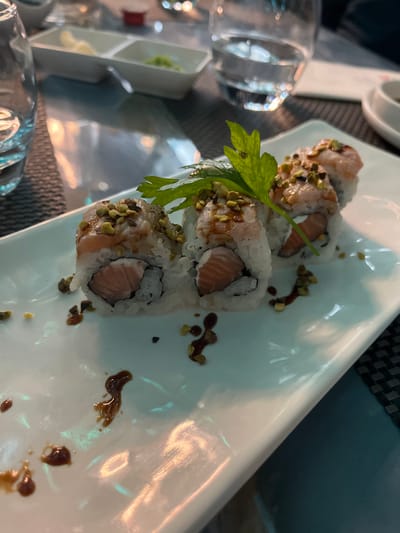 A close-up, slightly overhead shot captures four sushi rolls, likely uramaki, arranged on a white rectangular plate. The rolls feature rice on the outside, encasing what appears to be salmon and a lighter-colored ingredient, possibly cream cheese. They are generously topped with crushed green pistachios, creating a unique texture and color contrast. A dark, thick sauce, possibly a teriyaki or balsamic glaze, is drizzled artistically on the plate around the sushi, with some scattered pistachio crumbs. A fresh sprig of green parsley serves as a garnish on one of the rolls.

In the blurred background to the left, a drinking glass is visible, along with a small, compartmentalized white dish holding a green condiment (possibly wasabi) and another light-colored condiment. The sushi is placed on a dark, textured placemat or table surface. The scene depicts a dining experience, focusing on the beautifully presented sushi. The lighting suggests an indoor setting, possibly during the evening. There is no visible text in the image. The location is contextualized as Basiglio, Italy.