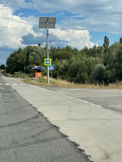 The image depicts a roadside scene in Pyrizhky, Ukraine, on a sunny day with a cloudy sky. The foreground shows a paved road, transitioning from asphalt to a concrete shoulder on the right. In the middle ground, a pedestrian crossing is marked on the concrete shoulder. Adjacent to the crossing is a bus stop with a small brick building and a shelter. A lamppost with a solar panel and a light fixture stands near the bus stop. Two pedestrian crossing signs are mounted on the lamppost. Behind the bus stop and road, dense green trees form a thick line. The overall environment suggests a rural or suburban area with public transportation access. There are no people visible in the image.