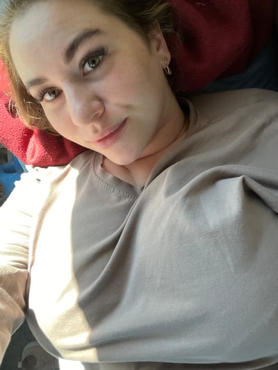 A close-up selfie shows a young Caucasian woman with light skin, brown hair, and green eyes. She is lying down, with her head resting on a red cushion and her body angled slightly away from the camera. She is wearing a tan t-shirt. Her hair is partially covering her face. The lighting suggests an indoor setting with natural light coming from the right side of the frame. No specific location in Nizhnyaya Korsa, Russia, is identifiable from the image.