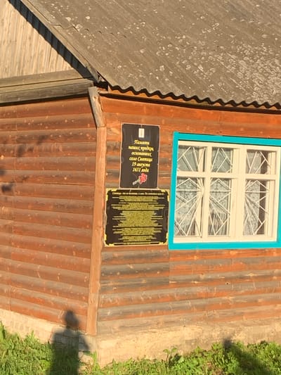 The image shows a wooden building with two plaques attached to its exterior wall. The building appears to be made of horizontal logs. A window with a blue frame is visible to the right of the plaques. The plaques are black with gold lettering and designs. The top plaque has text that appears to be in Russian, with a red rose image at the bottom. The bottom plaque also contains extensive Russian text. The scene is outdoors, with green grass and sunlight suggesting a daytime setting. A shadow of a person is visible on the ground near the base of the building. The plaques are indicative of a historical marker or dedication, possibly related to the founding or significant events in the village of Святица, Russia.