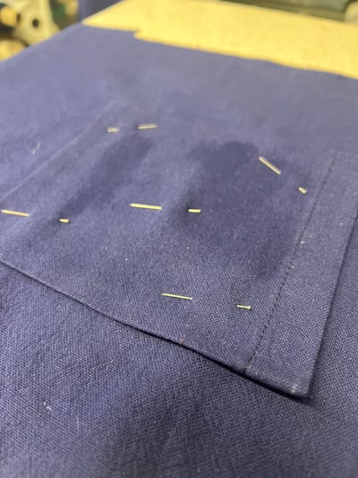 A close-up shot depicts a piece of navy blue fabric with a pocket sewn onto it. Several sewing pins are stuck into the fabric, securing the pocket in place. The fabric appears to be textured, possibly linen or a similar material. The setting is likely an indoor sewing or tailoring workspace, possibly within Kirklees, United Kingdom, although there is no explicit visual confirmation of this location. The primary subjects are the fabric and sewing pins, suggesting a garment construction or alteration process.