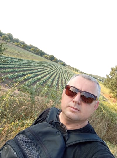 A man wearing sunglasses and a black t-shirt is taking a selfie in front of a field of crops. He is wearing a black backpack. The field is on a hillside, with rows of green plants stretching into the distance. Trees are visible on the surrounding hills. The sky is white and overcast. The location is Mereneshty, Moldova.