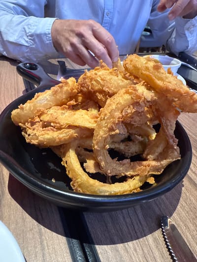 A close-up, high-angle shot captures a person reaching for a large platter of golden-brown fried food, likely onion rings, served in a black oval-shaped dish with a handle. The food appears crispy and is piled high in the center of the dish.

On the left side of the frame, a person is partially visible, wearing a light blue or white long-sleeved collared shirt. Their left hand is extended towards the platter, with fingers poised to pick up a piece of the fried food, indicating an active engagement with the meal.

The setting appears to be a restaurant or dining area, with a wooden table surface visible under the platter. Sunlight casts distinct shadows on the table, particularly from the dish and the serrated knife partially visible on the right side of the frame. A small portion of a white plate or bowl is also seen at the bottom right. The bright lighting suggests it is daytime. There are no discernible emotions from the partial view of the person, nor any visible text in the image. The location context for this scene is San Borja, Peru.