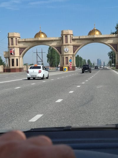 The media file depicts a street scene in Argun, Russia, oriented vertically. In the foreground on the right, a person's hand is visible, partially obscuring a dark object which might be a phone or camera. The main focus of the image is a wide asphalt road with white lane markings, curving gently into the distance.
Dominating the left side of the frame is a grand archway, constructed of beige stone with ornate carvings. Two golden domes top the structure, and smaller decorative elements are present. Mounted on the archway are several signs. Notably, a portrait of a smiling man in a red shirt is displayed on a lower section of the archway.
Several vehicles are on the road. A white hatchback, possibly a taxi due to the orange sign on its roof, is in the middle ground. Further down the road, a black SUV is visible, along with other vehicles that are less distinct. Overhead power lines and trees line the sides of the road. The sky is a clear, bright blue, suggesting a sunny day. The overall atmosphere is one of urban movement under clear skies.