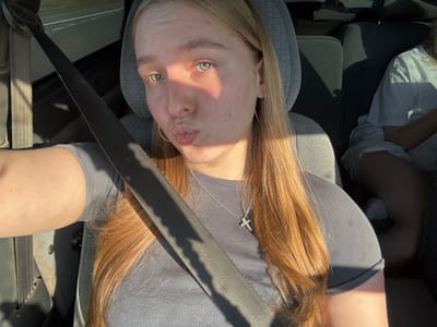 A young woman with blonde hair is sitting in the passenger seat of a car, wearing a grey t-shirt and a silver necklace with a cross pendant. Sunlight is illuminating her face, highlighting her green eyes and casting a shadow across her features. She is puckering her lips, as if for a kiss. A seatbelt is diagonally across her chest. In the background, another person is visible in the back seat of the car. The car appears to be on a road, possibly in a rural or suburban area given the sunlight and the hint of a road in the background. There is no specific indication of the location being in the city of Kukushtan, Russia, but the context of a car ride suggests it could be a journey within or near a populated area.