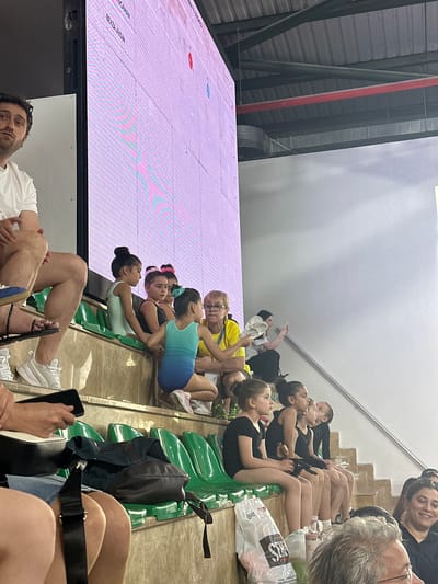 This image captures a scene from an event at the Sancaktepe, Turkey sports hall, likely a gymnastics competition, based on the attire of some participants. In the foreground, a man with dark hair and beard sits on a bench, looking up and to his left. To his right, several young girls in leotards are seated on green bleachers, some looking towards a large, illuminated screen displaying text and graphics, suggesting it's a scoreboard or information display. One girl in a turquoise leotard is closer to the viewer, interacting with an adult woman wearing a yellow top. In the background, more people are visible, some using mobile phones. The environment appears to be an indoor sports facility with tiered seating and overhead lighting structures. The focus is on the spectators and young athletes, with the large screen dominating the upper portion of the image.