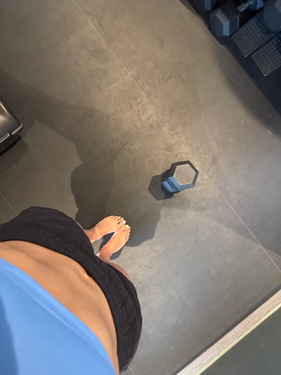 The image provides a high-angle view within a fitness facility in San Luis Potosí City, Mexico. The floor is composed of dark grey rubber matting, exhibiting scuffs and wear marks. A lone individual is partially depicted from the lower torso to the feet, standing barefoot. The person wears black athletic shorts and the lower portion of a light blue garment. Their toenails are painted a light yellow. A distinct, elongated shadow of the individual stretches across the floor to their left. A black hexagonal dumbbell rests on the floor to the right of the person's feet, also casting a shadow. In the upper right background, a rack holds multiple identical black hexagonal dumbbells and textured black plyometric steps. A dark, indeterminate piece of gym equipment is partially visible in the upper left corner. The lower right edge of the frame features a reflective strip, possibly a mirror or structural element.