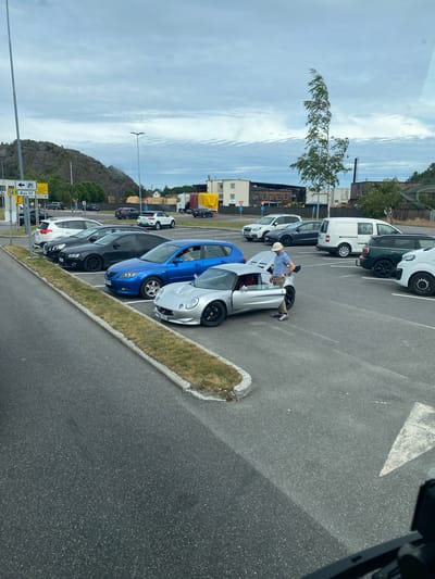 The image shows a parking lot in Larvik, Norway. A man wearing a light blue shirt, khaki pants, and a white hat is leaning into the open driver's side door of a silver sports car. The car appears to have its doors open, suggesting the man may be entering or exiting it, or perhaps showing it off. Several other cars are parked in a line next to the silver sports car, including a blue hatchback and a black sedan. Further down the parking lot, there are white vans and other vehicles. The scene takes place outdoors under a partly cloudy sky. In the background, there are buildings, a hill with some trees, and a road. There is also a yellow sign with Norwegian text, which includes "Øya 57" and directions to other locations. The overall impression is a casual daytime scene in a Norwegian town.