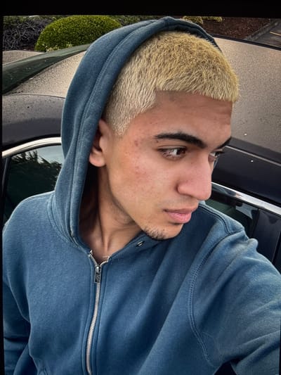 A young man is captured in a close-up, angled shot, wearing a hooded sweatshirt in a faded blue tone. He has light blonde, closely cropped hair. His skin tone is tan, and he has distinct eyebrows. The man is looking to his left, and his lips are softly closed. The background includes a glimpse of greenery and what appears to be a car exterior, suggesting an outdoor setting.