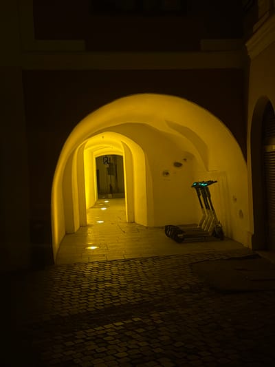 The image depicts a nighttime scene within a series of vaulted archways, characteristic of historic European architecture, specifically in the city of Sibiu, Romania. The foreground features a dark cobblestone ground, transitioning into a brightly illuminated, warm yellow passageway. This passageway is defined by multiple receding arches, each with light-colored, possibly cream or white, walls. The floor within the illuminated passage is paved with a smoother material, featuring at least three circular recessed floor lights that emit a focused, bright glow.
To the right, within the initial archway, three electric scooters are parked upright against the wall, their handlebars displaying small, active blue lights. A fourth electric scooter lies on its side nearby. The wall adjacent to the scooters shows minor surface irregularities and wear. To the far right, a dark, corrugated metal roll-down shutter is visible, indicating a closed entrance or storefront.
At the far end of the illuminated passage, a "P" (parking) sign mounted on a pole is discernible, suggesting an urban street beyond. No individuals are present within the frame. The overall scene is characterized by strong artificial illumination within the archway, contrasting with the darker, less lit areas in the immediate foreground and to the sides.