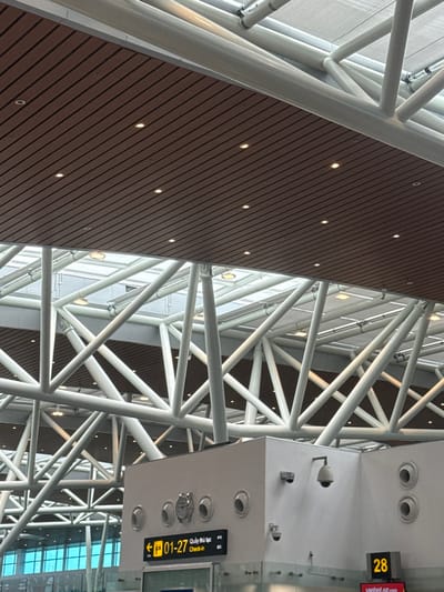 The image depicts the interior of a modern airport terminal, located in Hòa Cường Ward, Vietnam. The scene is dominated by a complex, multi-layered ceiling structure. The uppermost visible layer features an extensive white metal space frame or truss system, composed of numerous intersecting cylindrical beams forming a lattice. Portions of a lighter, possibly translucent ceiling or skylight are visible above this trusswork. Below the main truss structure, a section of brown, horizontally slatted false ceiling is present, uniformly embedded with multiple circular recessed spotlights.

In the lower right quadrant, a rectilinear white structural element, possibly a service counter or pillar, is visible. This element features several circular openings, likely vents, and is equipped with multiple surveillance cameras; specifically, one dome-shaped camera is mounted prominently, alongside smaller rectangular cameras. A black digital sign is affixed to this white structure, displaying yellow text that reads "<- 01-27 Quầy thủ tục Check-in," indicating directions to check-in counters. Below this, a partial red sign is visible with the number "28" and the text "vietjetair.com," suggesting an airline counter number and brand. No individuals or active interactions are present in the static frame.