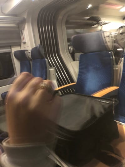 An interior shot of a train carriage. The blue patterned seats are visible. A black bag partially occupies the foreground, obscuring some of the scene. The train's ventilation system and wall panels are visible. In the foreground, a blurred hand is present, possibly holding or interacting with the bag. The lighting is soft and diffuse.