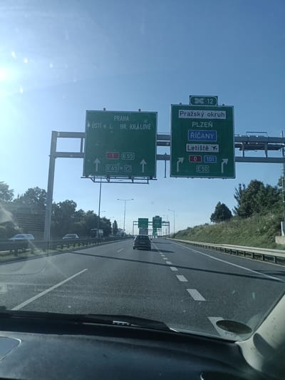 The image is a first-person perspective from inside a vehicle driving on a highway. Overhead green highway signs are visible. The sign on the left indicates directions to PRAHA, ÚSTÍ N. L., and HR. KRÁLOVÉ, with arrows pointing to the left and right. It also shows routes 1 E55 and E65. The sign on the right shows "Pražský okruh" (Prague Ring Road), PLZEŇ, ŘÍČANY, and Letiště (Airport), with route numbers 0, 101, and E50. The highway has multiple lanes and a clear sky overhead. Trees and grassy slopes line the side of the highway, and other vehicles are present on the road. The location appears to be a highway interchange on the outskirts of Prague, heading towards or within the area of Říčany, Czechia, given the signage.