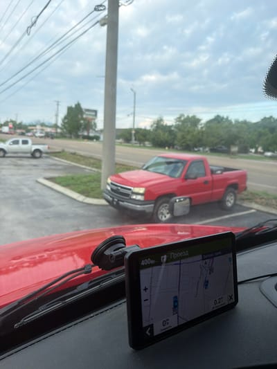 An eye-level medium shot captures the interior of a vehicle looking out a windshield at a red pickup truck parked in a lot. The pickup truck, a Chevrolet Silverado, is parked diagonally and facing left in the frame, with its driver's side door open and a side mirror extended. In the background, a white pickup truck is parked parallel to the curb on the left. A grey pole stands in the foreground between the camera and the red pickup truck, obscuring parts of the view. Beyond the parking lot, a road is visible with cars driving on it, and trees line the horizon under a cloudy sky. A GPS device is mounted on the vehicle's dashboard, displaying a map with navigation information. The image appears to have been taken in Chattanooga, Tennessee, given the displayed road names on the GPS such as "Tennessee Riverwalk".