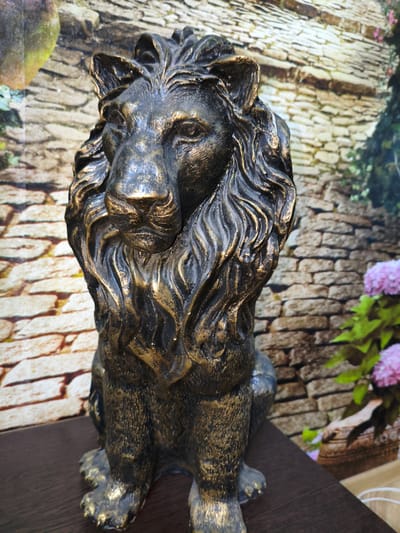 A bronze-colored statue of a lion with a detailed mane sits on a dark wooden surface. The statue is positioned facing slightly to the left, with its head tilted downwards. In the background, a printed wallpaper depicts a stone wall with a vine motif and pink flowers. The lighting is dim, with some highlights on the statue. The location is in Kotovsk, Russia.