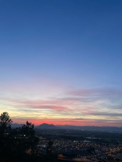 The image depicts a cityscape under a vibrant sunset sky, likely taken from a high vantage point in Rojales, Spain. The sky transitions from a deep blue on the left to streaks of pink, white, and yellow near the horizon. In the distance, mountains are silhouetted against the fading light. Below, a town is visible with numerous lights indicating nighttime activity. In the foreground, dark foliage suggests the presence of trees or bushes close to the viewing point. The composition includes natural elements and urban development, with the sky being the dominant feature.