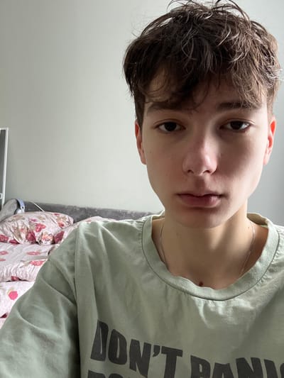 The image displays a headshot of a young individual, possibly a teenager, positioned indoors in what is identified as the city of Zainsk, Russia. The person, with light skin and short, disheveled dark brown hair, faces directly towards the camera, maintaining a neutral expression with eyes open. They are wearing a light sage green short-sleeved t-shirt featuring dark grey text, partially visible, which reads "DON'T PANIC". A thin silver chain necklace is visible around the neck.
The background consists primarily of a plain light grey wall. To the left and behind the person, a bed is visible with a dark grey upholstered headboard. The bedding includes a light-colored duvet or sheet adorned with a pink and red floral pattern. A pair of light blue over-ear headphones rests on the headboard, partially obscured, to the left of the person's head. The lighting is even, suggesting an interior light source or diffused natural light.