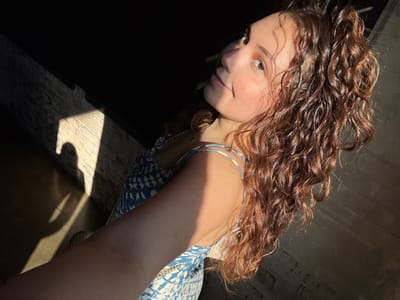 A woman with curly, brown hair looks at the camera in a close-up shot. She has fair skin and is wearing a blue and white patterned top. Her face is turned slightly to the side, and her eyes are open. She has a silver bracelet on her arm and a necklace around her neck. Part of the background is light-colored brick. Sunlight streams in from the side.