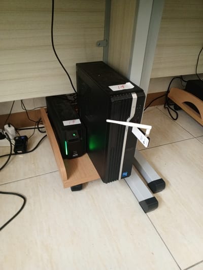 In a room with light-colored tiled flooring and a light-wood paneled wall, a black Acer desktop computer tower is positioned on a gray stand. To the left of the computer, on a light-wood shelf, sits a black APC uninterruptible power supply (UPS). A white antenna, likely a Wi-Fi adapter, is plugged into the front of the computer. Several black cables are visible, connecting the devices to power sources and possibly to each other. A white label with red numbering "14" is affixed to the top of the computer tower, and another similar label is on the UPS. A small black power adapter is situated on the floor to the left of the UPS. The scene is clean and organized, suggesting a functional office or computer station. There are no people or animals present in the image. The location in Chutula, Mozambique is not indicated by any visual cues within the image.