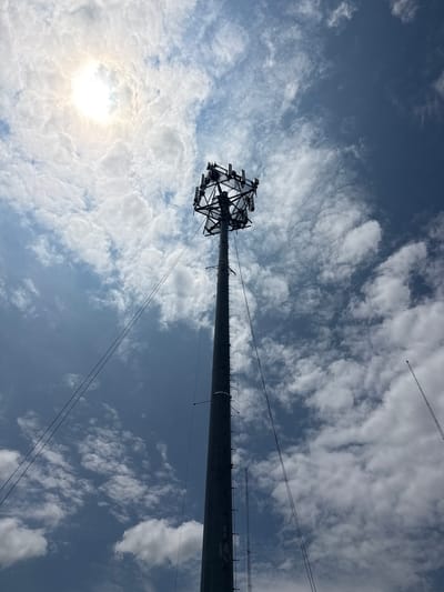 This is a low-angle, wide shot of a large tower against a blue sky with scattered white clouds. The sun is visible on the left side of the image, peeking through the clouds, suggesting it is daytime. The tower is a prominent object, its upper section featuring a complex lattice structure of metal beams. Cables extend from the tower and stretch across the frame. In the distance, high in the sky, faint contrails of an airplane can be seen. The overall impression is of a bright, clear day with industrial infrastructure against a natural backdrop. There are no people visible in the image.