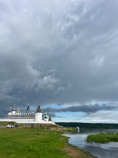 The image depicts a view of the Makarievsky Monastery in Makar'evo, Russia, situated on a hill overlooking a wide river. The monastery complex features several white walls, numerous onion-domed churches with gold spires, and a prominent white cylindrical tower with a dark cap. Adjacent to the monastery, a paved road runs along its base, with several vehicles, including a white SUV and a darker van, parked or driving on it. A few individuals are visible near the road and on the monastery grounds.
The foreground of the image is dominated by a lush green grassy field, leading down to the sandy bank of the river. The river itself is calm, with ripples reflecting the overcast sky and the dark green tree line on the opposite bank. A small white boat is docked on the far side of the river, near the forest.
The sky is filled with dramatic, dark grey storm clouds, creating a moody atmosphere. Patches of lighter sky and blue are visible in the distance, suggesting the storm is either approaching or moving away. The lighting is diffused due to the cloud cover. The overall scene captures a moment of natural beauty and historical architecture under a dynamic sky.