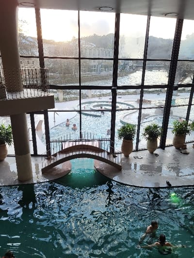 View of a large window overlooking a pool. Outside, there is another body of water visible with multiple people in it. A small part of the pool is visible, with some people swimming in it. Near the window, there are planters with greenery in them. The sun is setting in the distance, creating a warm glow.