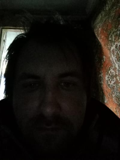 A dimly lit close-up photograph features a person with dark hair and facial hair. The background includes a partially visible patterned tapestry in shades of orange and green on the right side, and a blurred frame of a window or doorway on the left side. The location appears to be indoors, potentially in a private residence. No specific information is available to indicate the location is in Parabel, Russia.