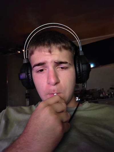 A close-up selfie shows a young man wearing black over-ear headphones with a white illuminated headband. He has short, brown hair and is looking downwards, with his right hand raised to his mouth, holding a white object, possibly a cigarette or a lollipop. His expression is neutral. He is wearing a greyish-green t-shirt. The background is dark, suggesting an indoor setting, possibly a bedroom or gaming setup, with some blurred lights and shelves visible in the distance. The overall lighting is dim, with some colorful illumination from the headphones and the background. The man is positioned in the foreground, with the background out of focus. There is no indication of the location being Aleksandrovo, Bulgaria.