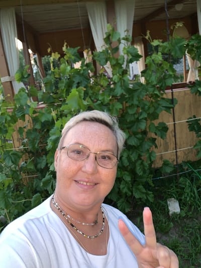 A medium close-up selfie of a woman with short blonde hair and freckles, wearing glasses and a white t-shirt, is taken outdoors. She is smiling and holding up two fingers in a peace sign. Behind her, a green vine with large leaves is visible, along with a wooden structure with white curtains and a yellow wall. The background appears to be a residential area, possibly in the city of Суроватиха, Russia. The lighting suggests it is daytime.