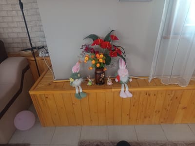 This is an indoor scene depicting a wooden chest against a light-colored wall. On top of the chest, there is a vase filled with red anthurium flowers and smaller yellow flowers. Two gnome-like figures with pink ears and beards are placed on either side of the vase. To the left of the gnome, there are two small bunny figures and a decorative vase. To the far left, a pink balloon is visible on the floor. A floor lamp with a black pole stands to the left of the chest, against a brick-patterned wall. The right side of the image shows a sheer white curtain. This image is located in Radishevo, Bulgaria.