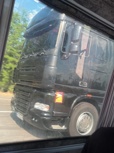 The image shows a close-up view of the front and side of a black truck, possibly a semi-truck or a large cargo vehicle. The perspective is from inside another vehicle, looking out through a window, as indicated by the reflections and the framing of the image. The truck is black and appears to be in motion, with the wheel at the front right of the truck slightly blurred, suggesting movement.

Notable details include a yellow and red warning sticker on the front of the truck, near the headlight. The sticker has text and a graphic, though the text is too blurry to read. A headlight is visible, and part of the truck's grille. The background seen through the truck's windows and reflected on its surface shows green trees and a bright sky, suggesting it is daytime and the weather is clear. The orientation of the image is unusual, with the truck appearing upside down, which might be due to the camera's positioning or a post-processing effect.

The scene is likely set on a road, given the context of being inside a moving vehicle and the appearance of the truck. There are no people clearly visible. The overall impression is a snapshot of a vehicle encountered during travel.