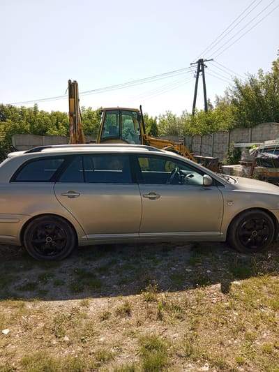A beige station wagon is parked on a dirt and grass lot in Leoncin, Poland. The car is facing right, and its side is visible. The car has black rims and tinted windows. In the background, there is a yellow excavator, a concrete fence, trees, and a utility pole with power lines. The sky is clear and blue.