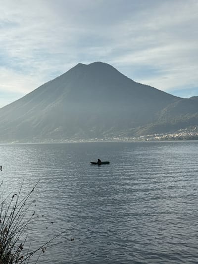 The image displays a wide view of a large body of water, likely Lake Atitlán, set against a mountainous backdrop in San Pablo La Laguna, Guatemala. The sky is light blue with scattered, wispy white clouds. A prominent, dark blue-grey, conical mountain, covered with dense vegetation, dominates the background, rising from the distant shoreline. Atmospheric haze subtly softens the mountain's contours. Along the base of the mountain on the far right, a cluster of small structures indicates a distant human settlement. The lake surface is dark blue-grey with numerous ripples, suggesting gentle movement. In the mid-ground, a single, dark-colored, small watercraft, resembling a canoe or cayuco, floats on the water. One individual is seated or kneeling within this boat, presented as a dark silhouette, facing slightly to the left. No specific actions or equipment (e.g., paddles, fishing gear) are discernible for the person or boat. In the lower-left foreground, several tall, thin, reedy plants extend upwards, partially obscuring the immediate water's edge.