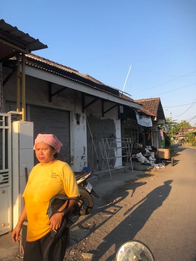 In the city of Kalidawir, Indonesia, a woman wearing a pink bandana and a yellow t-shirt stands by the roadside. The t-shirt has the text "BEE BRAND 1000" printed on it. She is holding a dark bag over her shoulder. The background shows a building with a tiled roof and a closed metal shutter. In front of the building, there is a clothes drying rack and various items piled on the ground. A motorcycle is parked behind the woman. The scene appears to be a street in a residential or commercial area, with buildings and trees visible along the road. The lighting suggests it is either early morning or late afternoon, with long shadows cast on the ground.