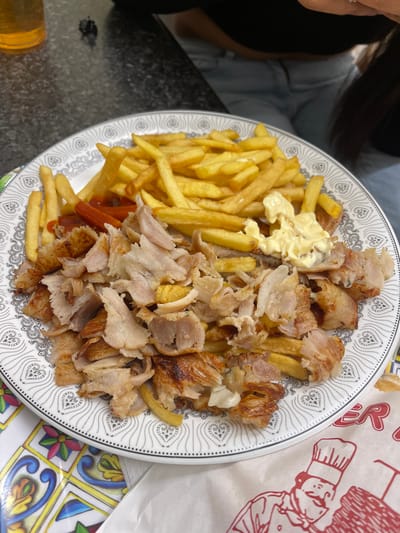 This close-up image captures a large, casual meal served on a plate in an indoor setting, likely in Sondrio, Italy. The central subject is a generous portion of food, predominantly comprising golden-brown french fries and numerous strips of shaved meat, characteristic of döner kebab (appearing to be chicken or turkey). A dollop of creamy white sauce, possibly mayonnaise or garlic sauce, is visible on a pile of fries, with traces of red ketchup also present beneath some of the fries.
The food is presented on a white, circular plate featuring a distinct black and white patterned border composed of repeating heart-like or teardrop motifs. The plate rests on a dark, possibly granite or marble, countertop or table surface. To the right, a vibrant section of a table or placemat with colorful, geometric tile patterns adds a contrasting visual element.
A person is partially visible in the upper left of the frame, showing a dark sleeve or garment, part of a hand with a ring, and dark hair, suggesting they are seated at the table. In the background, a hint of light blue clothing indicates the presence of another individual. At the bottom left, a golden-colored glass, possibly containing a beverage, is partially visible.
To the right of the plate, a crumpled white paper bag or wrapper is visible. It features red text, with the letters "ER" clearly discernible, and a stylized illustration of a chef's head and hat. This packaging suggests the meal may be from a takeaway or a casual eatery. The scene depicts a typical mealtime, such as lunch or dinner, in a relaxed environment.