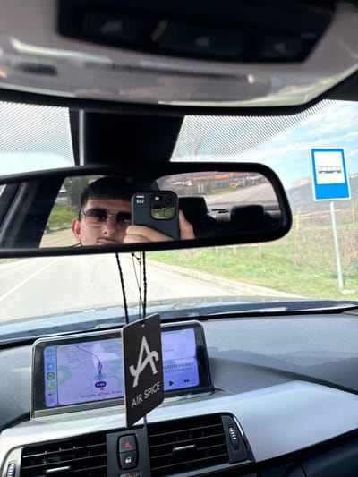 The image depicts the interior of a vehicle, looking towards the dashboard and the front windshield. A male individual, wearing sunglasses, is reflected in the rear-view mirror, holding a dark-colored smartphone with a case displaying the Bulgarian flag. The smartphone's screen shows Cyrillic text "ЯЗВ". An "AIR SPICE" air freshener hangs from the rear-view mirror stem. The vehicle's dashboard features a textured light grey trim and a central infotainment screen. This screen displays a navigation map indicating a route with an estimated arrival time of 14:11, 47 minutes, and a distance of 53 kilometers. Visible on the map are Cyrillic place names, including "Летница Драгановци" (Letnitsa Draganovtsi) and "Яворец" (Yavorets), confirming the location in the Draganovtsi region of Bulgaria. The current time on the screen is 13:24. Through the windshield, a blue square bus stop sign with a white bus icon is visible on the right side of the road, set against a backdrop of green grassy terrain and distant hills under a partly cloudy sky. The road appears to be asphalt, leading through a semi-rural environment.