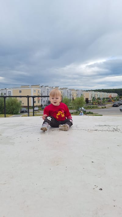 In Myza (Inshinskii S/O), Russia, a young child with blonde hair is sitting on a light-colored, flat, outdoor surface. The child is wearing a red long-sleeved shirt with a graphic print and dark pants with a floral pattern. They are looking towards the viewer with a joyful expression. In the background, a row of apartment buildings with light-colored facades and dark roofs is visible. The sky is overcast with gray clouds. A metal fence runs horizontally across the mid-ground, separating the sitting area from a road and a playground. Several cars are parked on the side of the road, and some trees are present.