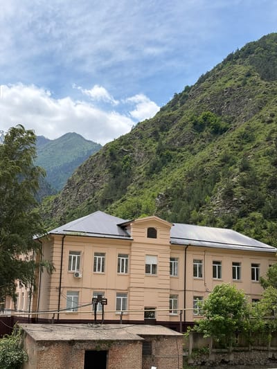 This is a full-shot, eye-level, outdoor photograph taken in Mizur, Russia. The scene depicts a multi-story building with a beige facade, a dark metal roof, and numerous white-framed windows. The building is situated in a valley, with a steep, green, rocky mountainside rising behind it. Lush trees are visible on the left and right sides of the building, and in the foreground, there is a lower, brick structure with a flat roof. The sky is partly cloudy with a blue hue. The photograph is in landscape orientation.