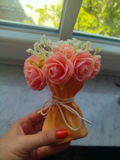 A hand with bright orange nail polish is holding a small, decorative vase filled with artificial pink roses and tiny white flowers. The vase is a bright orange color and is tied with a thin, off-white ribbon. The background is blurred, showing a window with bright sunlight filtering through green foliage. The scene suggests a bright, sunny day, possibly in the morning or afternoon. The overall impression is cheerful and decorative, with the focus on the artificial floral arrangement. The location context provided is Offenbach am Main, Germany, but there are no specific visual cues in the image to confirm this. There is no visible text in the image.