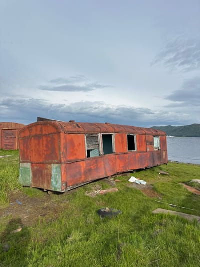 The image captures a desolate scene in Bolshaya Rechka, Russia, featuring an abandoned, rusty red train car positioned on grassy terrain next to a body of water. The train car, showing signs of decay with peeling paint and broken windows, rests on wooden supports. In the background, a tranquil body of water is visible, with a distant green, mountainous shoreline under a cloudy sky. Another abandoned structure, possibly another train car, is partially visible to the left of the main subject. The immediate surroundings are covered in lush green grass, interspersed with scattered debris, including a discarded tire and pieces of wood. The overall atmosphere is one of abandonment and natural reclamation.