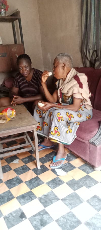 An interior scene in Udi, Nigeria, features two women. The woman on the left is a younger adult, dressed in a black and red striped short-sleeved top, seated on a low surface or floor, observing the other woman. Her hands are clasped in front of her. The woman on the right is an older adult with short, grey hair, wearing a patterned dress (white with yellow, red, and black swirls) and a beige and maroon striped shawl. She is seated on a red upholstered couch, actively eating a white food item with her right hand while holding another piece in her left. She wears light blue flip-flops.
Between the two individuals is a light brown wooden table. On the table, a crumpled red, white, and yellow plastic wrapper is visible. The floor is tiled in a black and orange/light brown checkerboard pattern. In the background, a dark brown wooden shelving unit with items, including red artificial flowers, is present behind the woman on the left. To the right, a striped curtain (grey and white vertical stripes) is partially visible. The walls are a plain light beige.