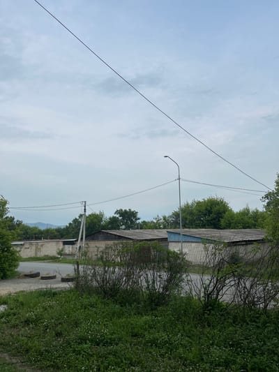 The image depicts a street scene in Smolyaninovo, Russia, under a cloudy sky. In the foreground, lush green grass and bushes dominate, with small white flowers scattered throughout. A paved road runs through the midground, bordered by an array of garages or industrial buildings constructed from light-colored brick with corrugated metal roofs. A tall utility pole with overhead wires stands to the left of the road, and a street lamp is visible further down. In the background, a line of trees forms a dense green border, with distant hills or mountains visible on the horizon. Two large tires filled with sand are positioned on the side of the road near the utility pole, serving as impromptu planters. The overall impression is one of a utilitarian, somewhat unkempt, semi-rural industrial area.