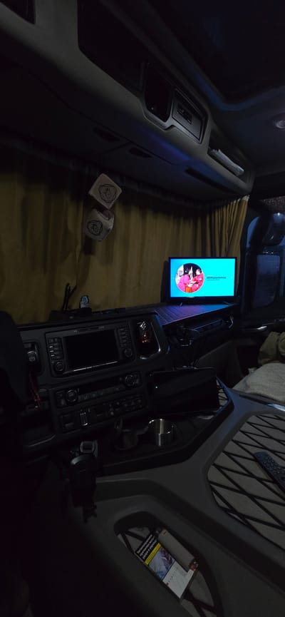 This image depicts the interior of a truck cab at night, likely in Épila, Spain. The focus is on the dashboard area and the rear sleeping compartment. A television is mounted on a shelf, displaying a cartoon with Russian text. On the dashboard, there's a complex control panel with various buttons and a screen, and to the right, a silver cup holder. Further back, a tan curtain lines the sleeping area. Two cubic decorations, possibly fuzzy dice, hang from the ceiling. In the lower right corner, a pack of cigarettes and a lighter are visible in a storage compartment. The overall lighting is dim, suggesting nighttime.